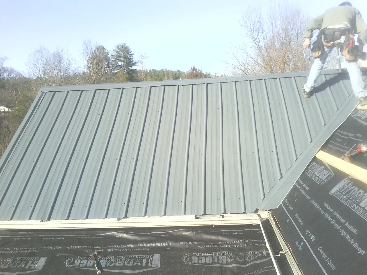Crew installing dark green standing seam panels over synthetic underlayment in Manteo
