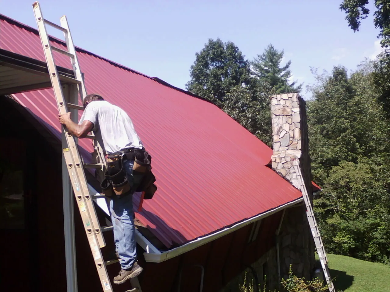 Roofer on a ladder beside a red metal roof and stone chimney — Soffit & Fascia Repair by Roofing Brothers in Manteo